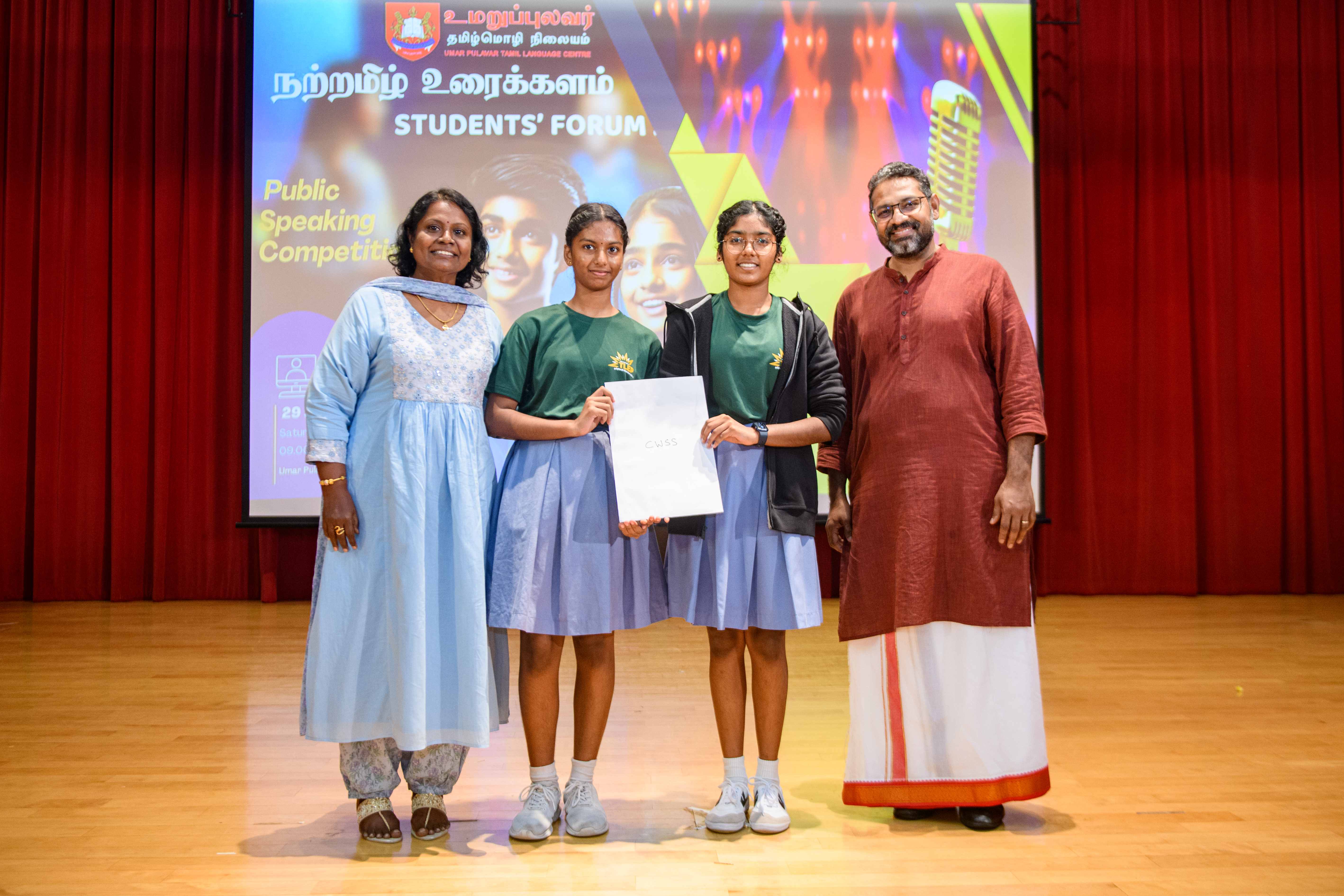 Four people on stage pose with two schoolgirls holding a certificate; backdrop reads Students’ Forum.
