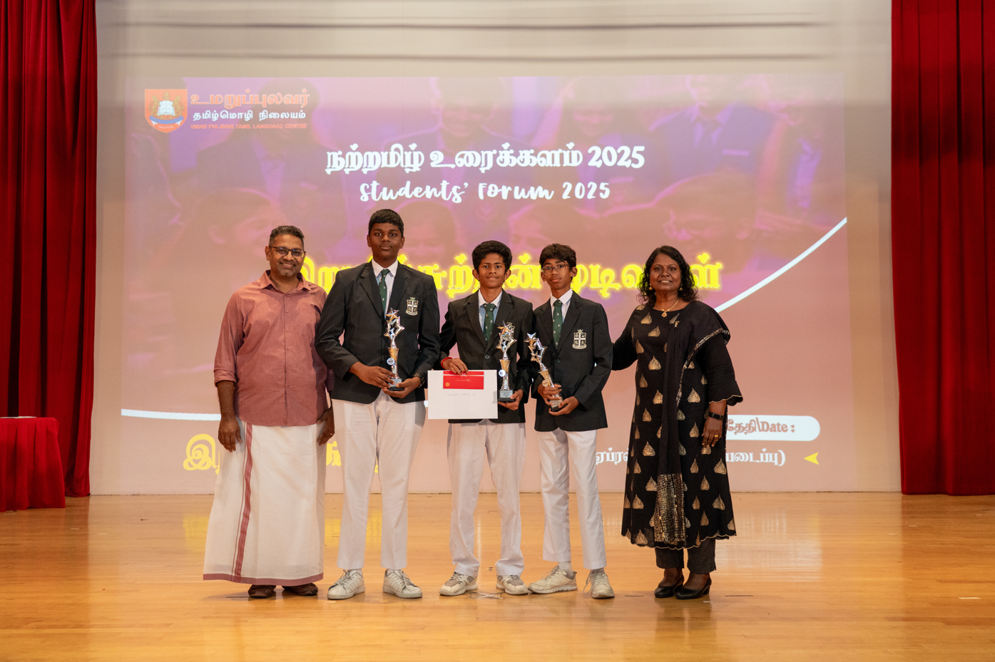 Five people on stage; three students in blazers hold trophies and a certificate before a Students’ Forum 2025 screen.