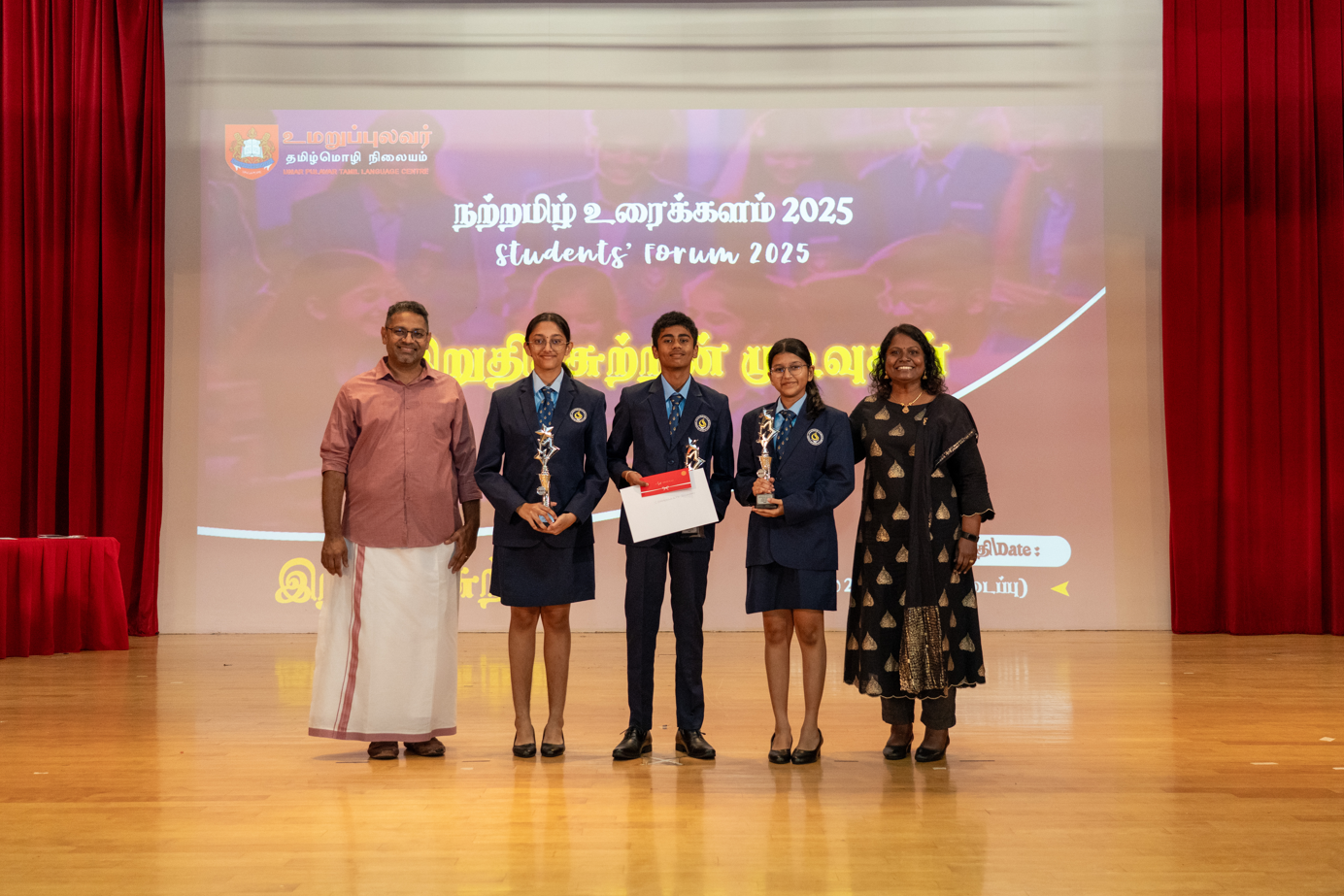 Five people on stage; three students in uniforms hold trophies and a certificate before a Students Forum 2025 screen.