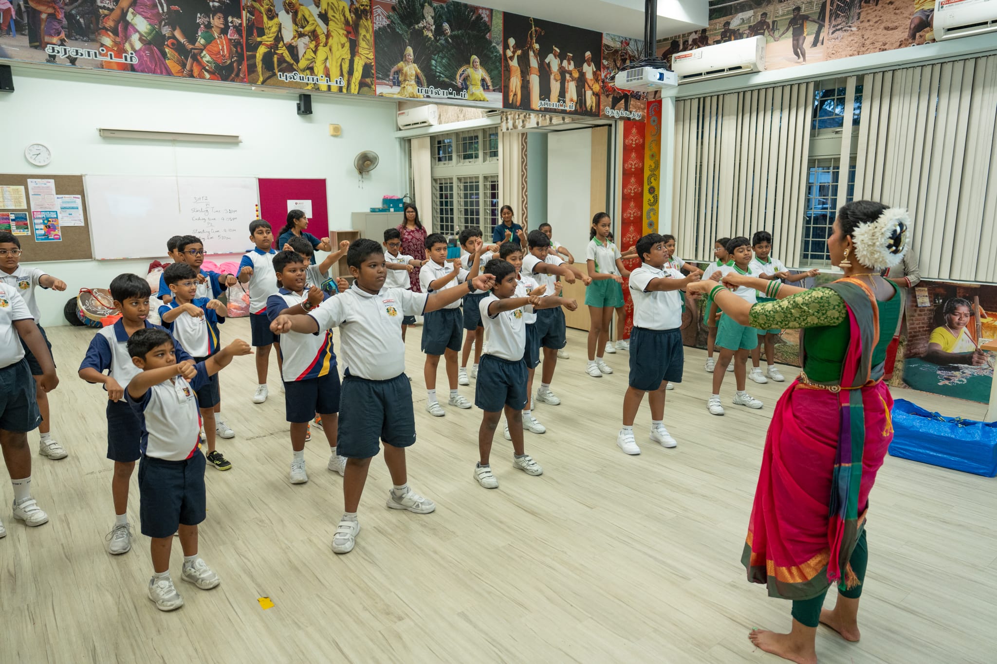 Teacher in sari leads schoolchildren in uniforms doing arm poses during a dance class in a bright room