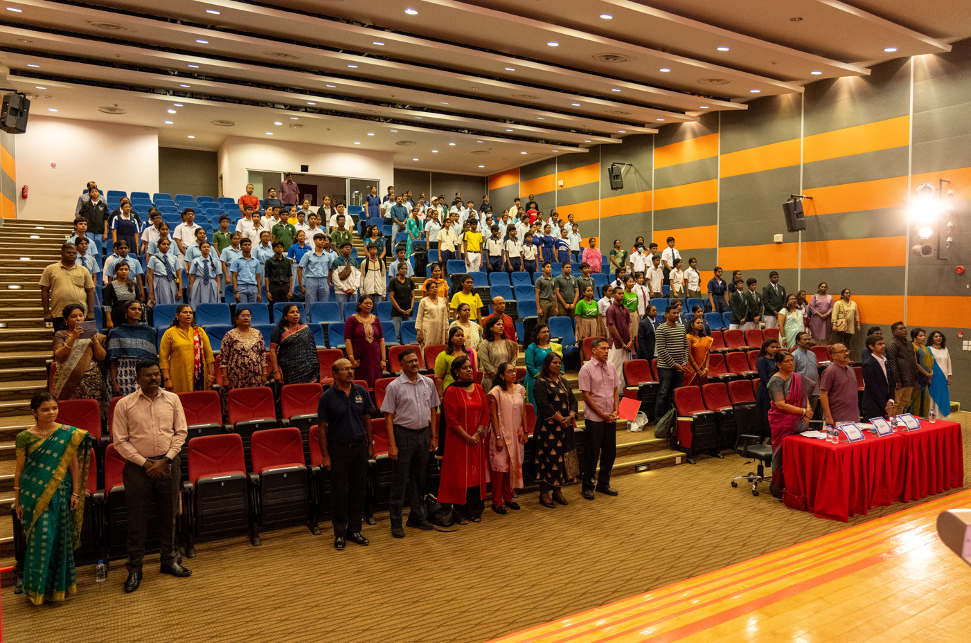 Crowd of students and adults standing in an auditorium with blue seats; red table with nameplates at front.