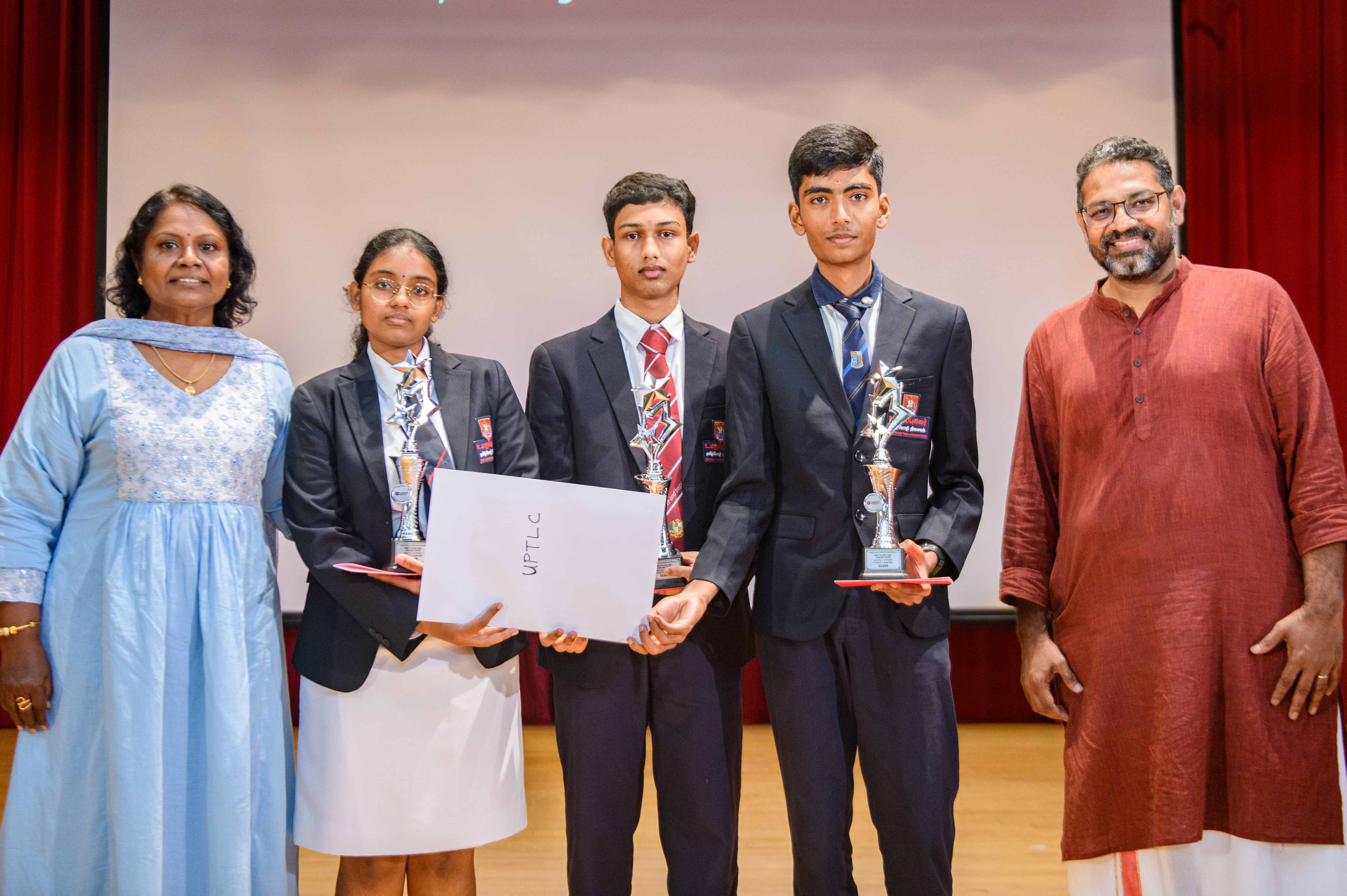 Five people on a stage; three students in blazers hold trophies and a certificate, flanked by two adults.