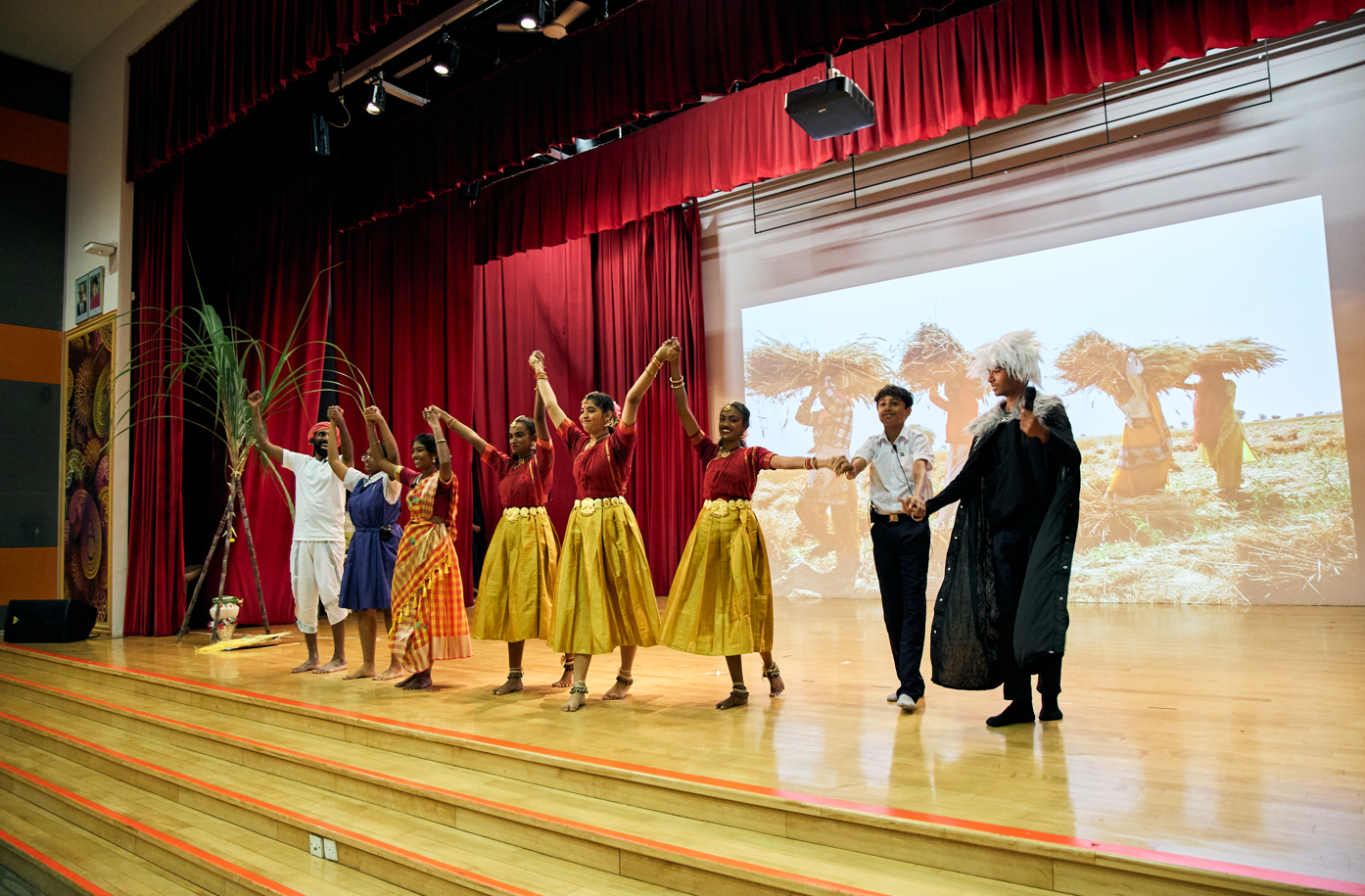 Performers hold hands and bow on a stage with red curtains, while a field scene is projected behind.