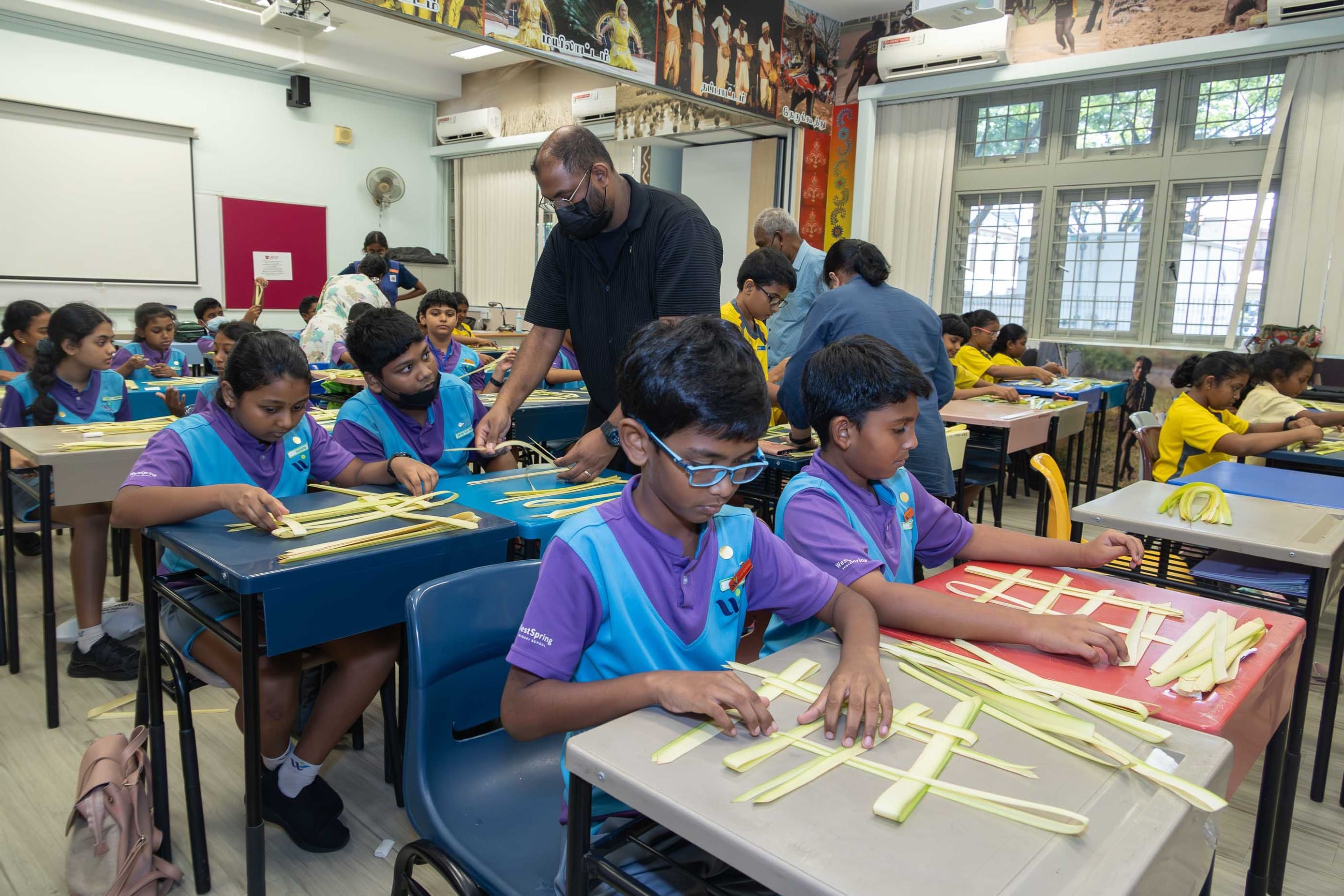 Teacher assists schoolchildren weaving pale palm strips at desks in a classroom with large windows.