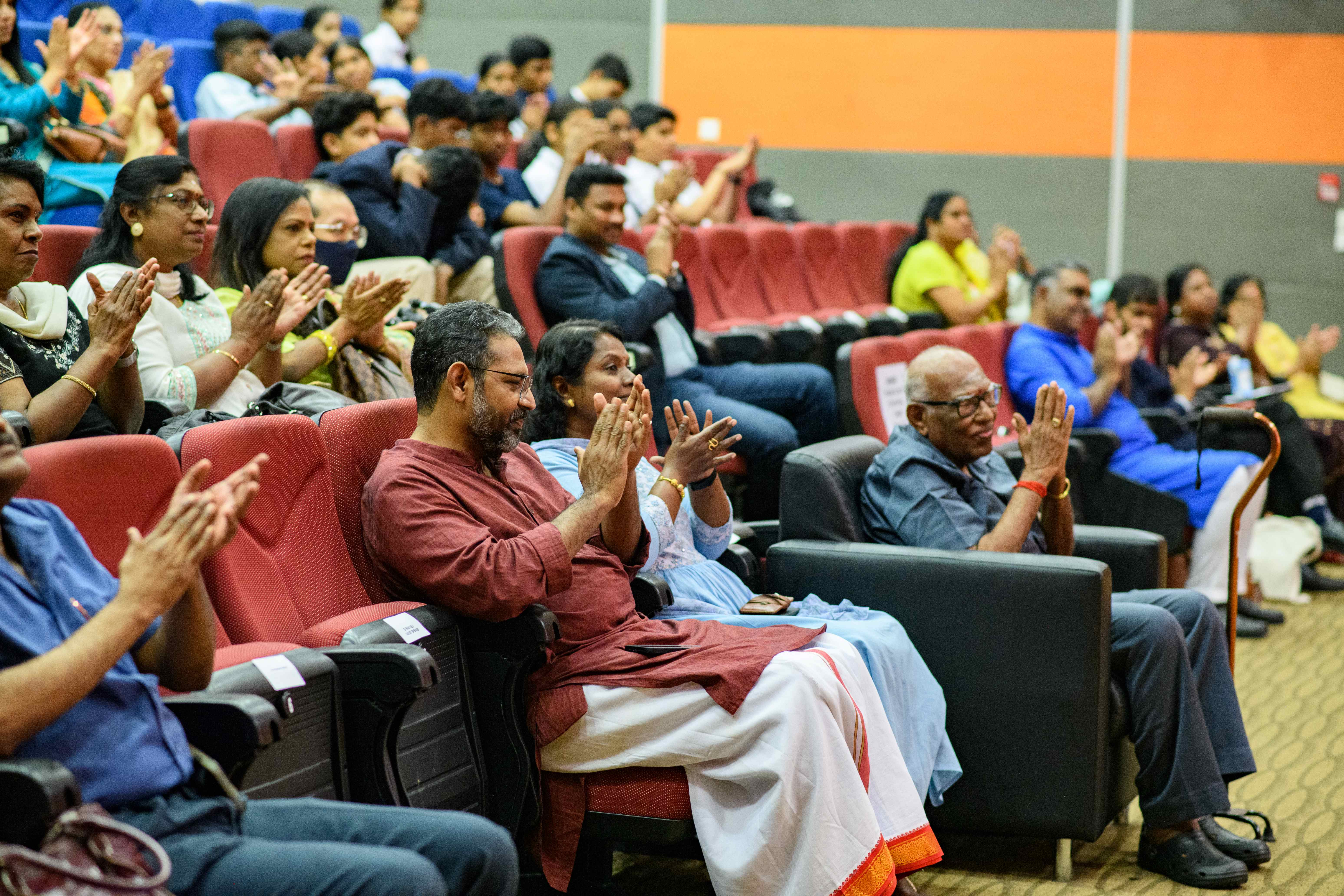 Audience seated in a theater applauds, with people in red seats and a few in front-row armchairs.