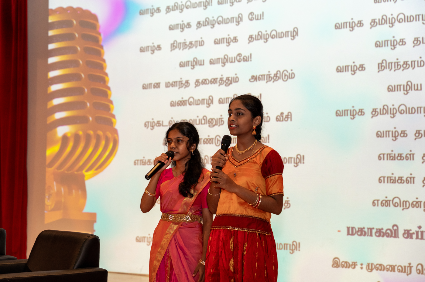 Two girls in colorful traditional dress hold microphones on stage before Tamil text and a large mic graphic