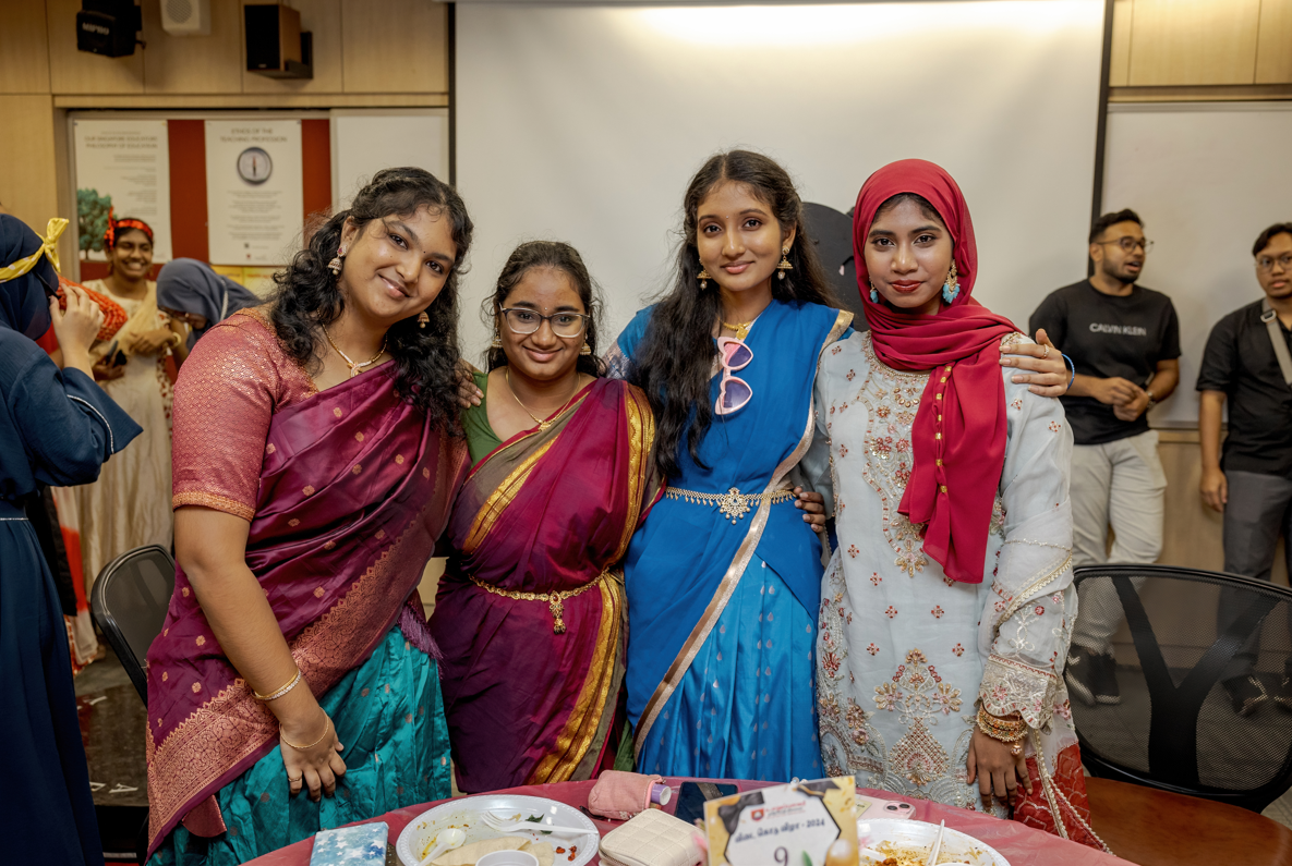 Four women pose with arms around each other in colorful traditional outfits at an indoor gathering.