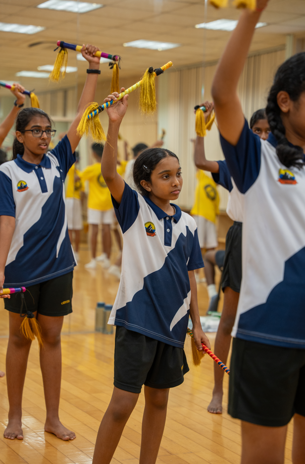 Girls in sports uniforms practice with colorful sticks and yellow tassels in a mirrored studio.