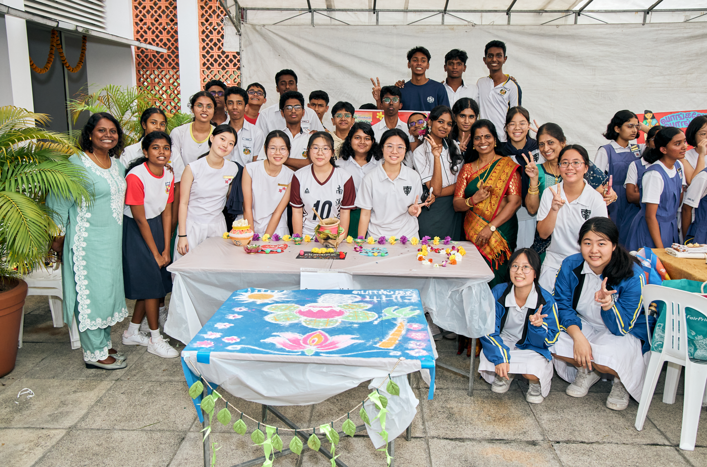 Group of students and teachers posing behind decorated tables, many making peace signs, with a rangoli board in front.