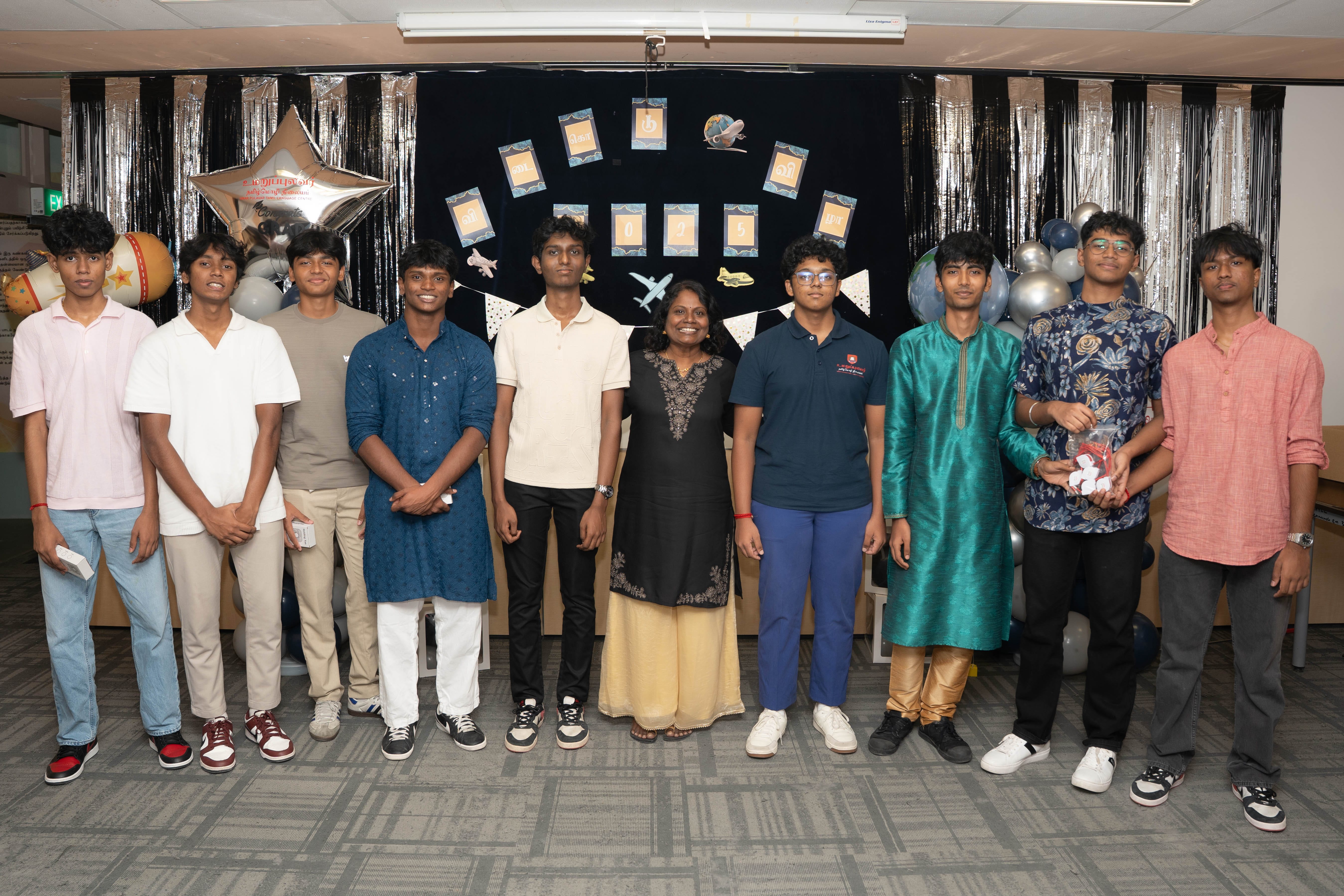 Group of 10 posing indoors before a decorated backdrop with balloons, tinsel curtains, and hanging cards.