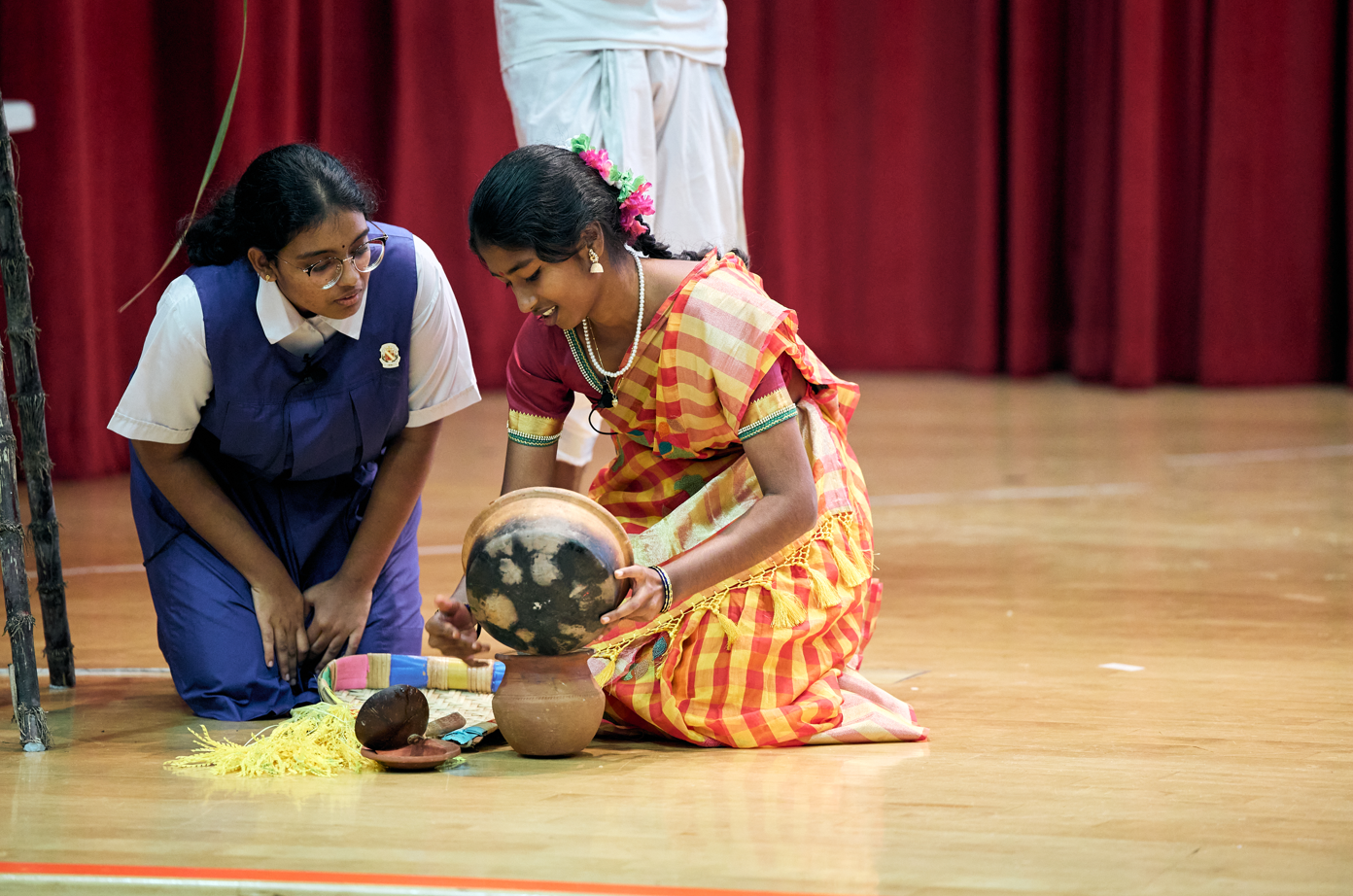 Two girls kneel on a stage; one in a sari pours from a pot into a bowl while the other watches.