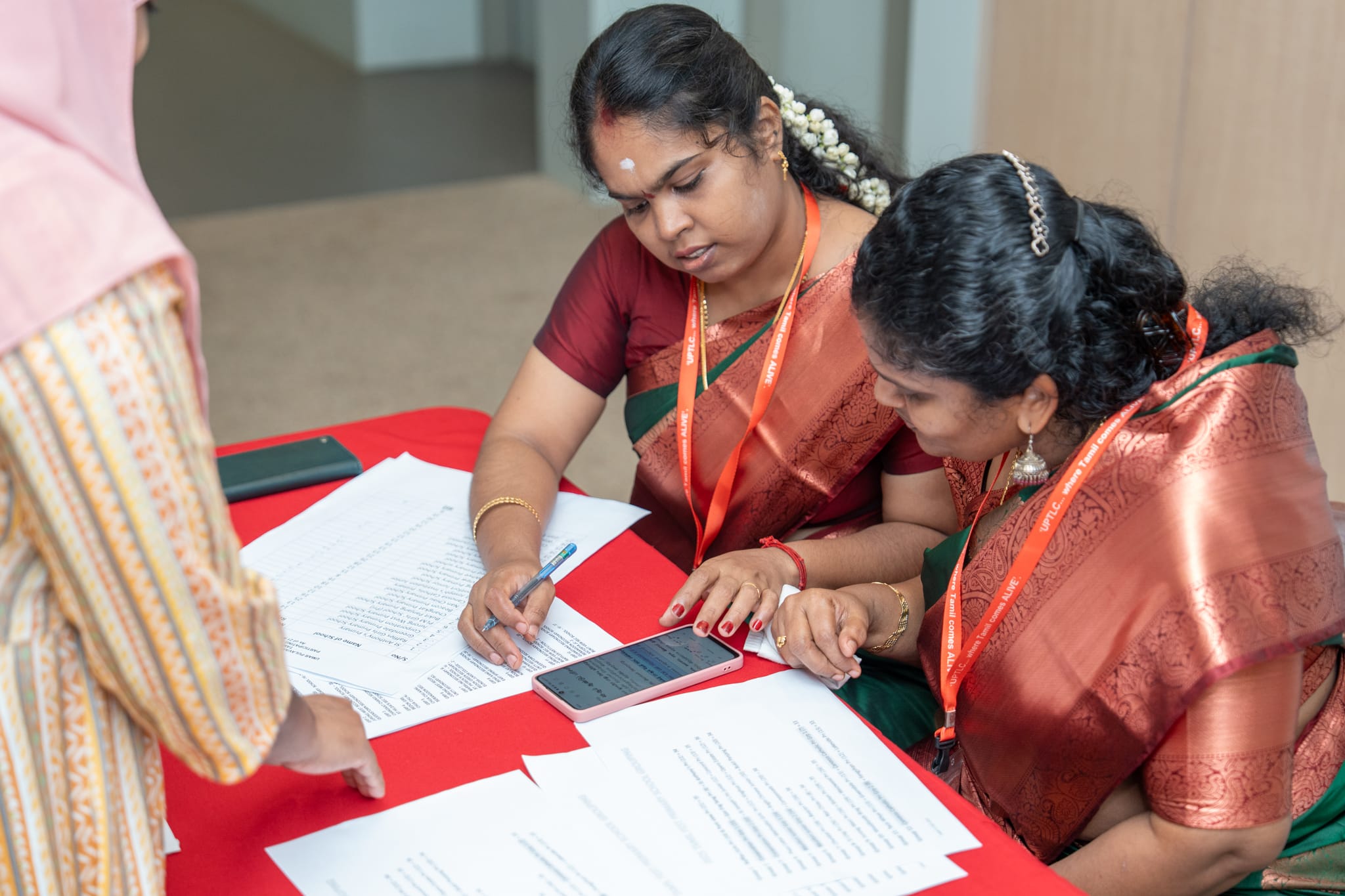 Two women in sarees review papers and a phone at a red table while a third person stands nearby.