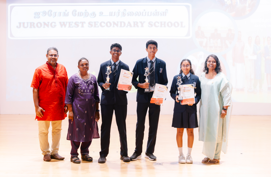 Six people on stage; three students in uniforms hold trophies and certificates under a school banner.