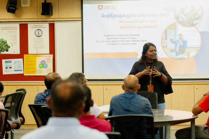 Woman speaks to seated audience in classroom, with projection screen showing Tamil text behind her.
