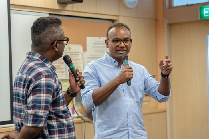 Two men with microphones speak indoors; one in a blue shirt gestures while the other in plaid listens.
