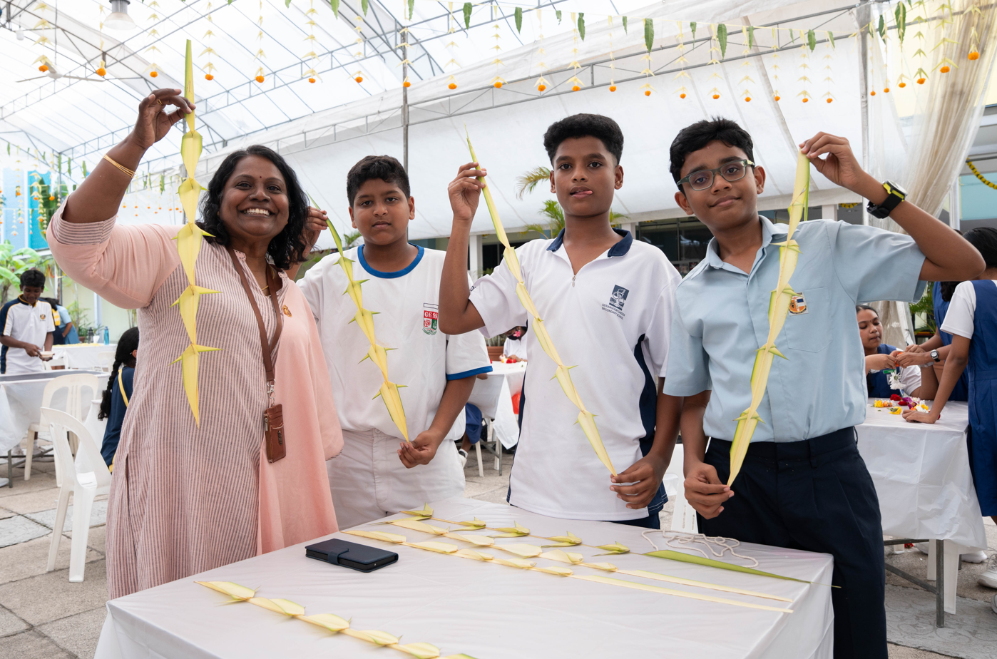 Woman and three boys hold long woven palm leaves under a canopy, with more palm pieces on a table.