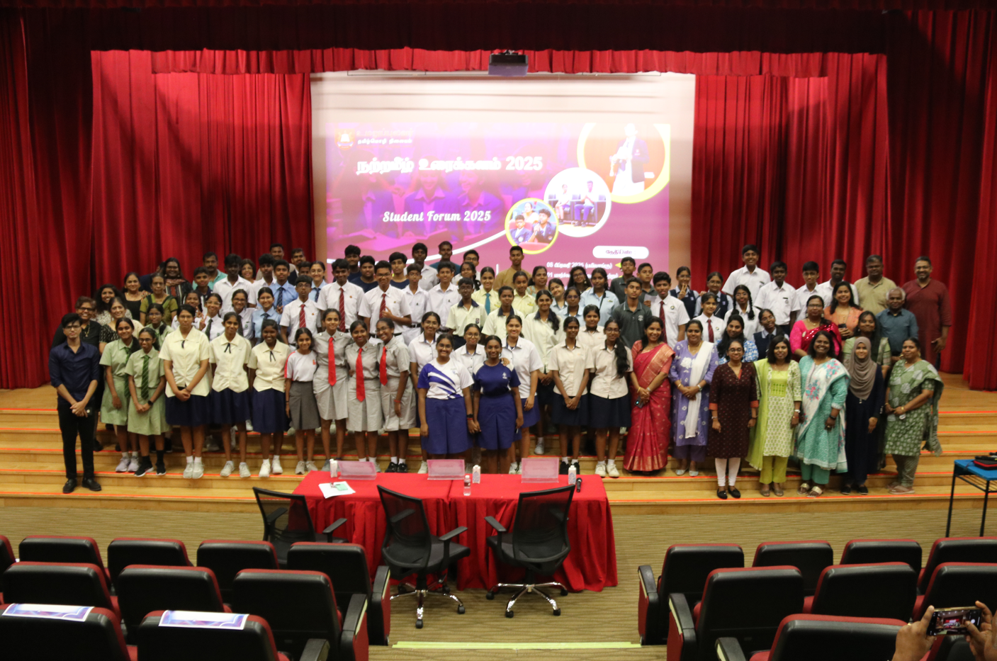 Large group of students and staff posing on stage with red curtains and a projected Student Forum 2025 banner.