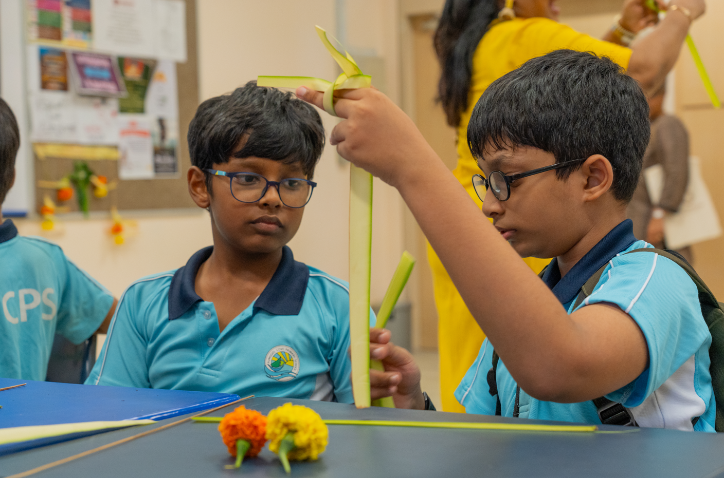 Two boys in blue school uniforms and glasses weave green leaf strips at a table with small flowers.