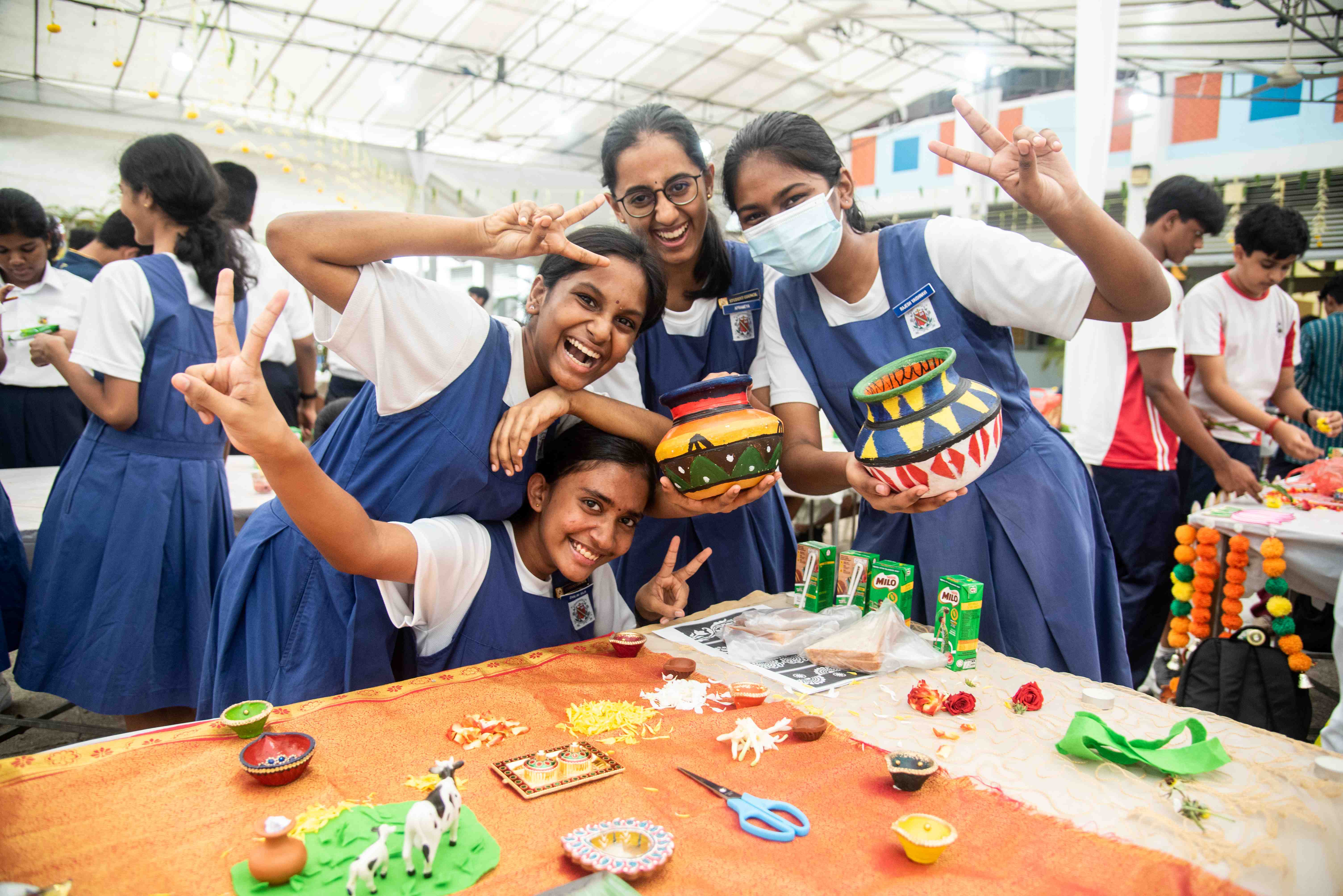 Four schoolgirls in blue uniforms smile and flash peace signs, holding painted pots at a table.