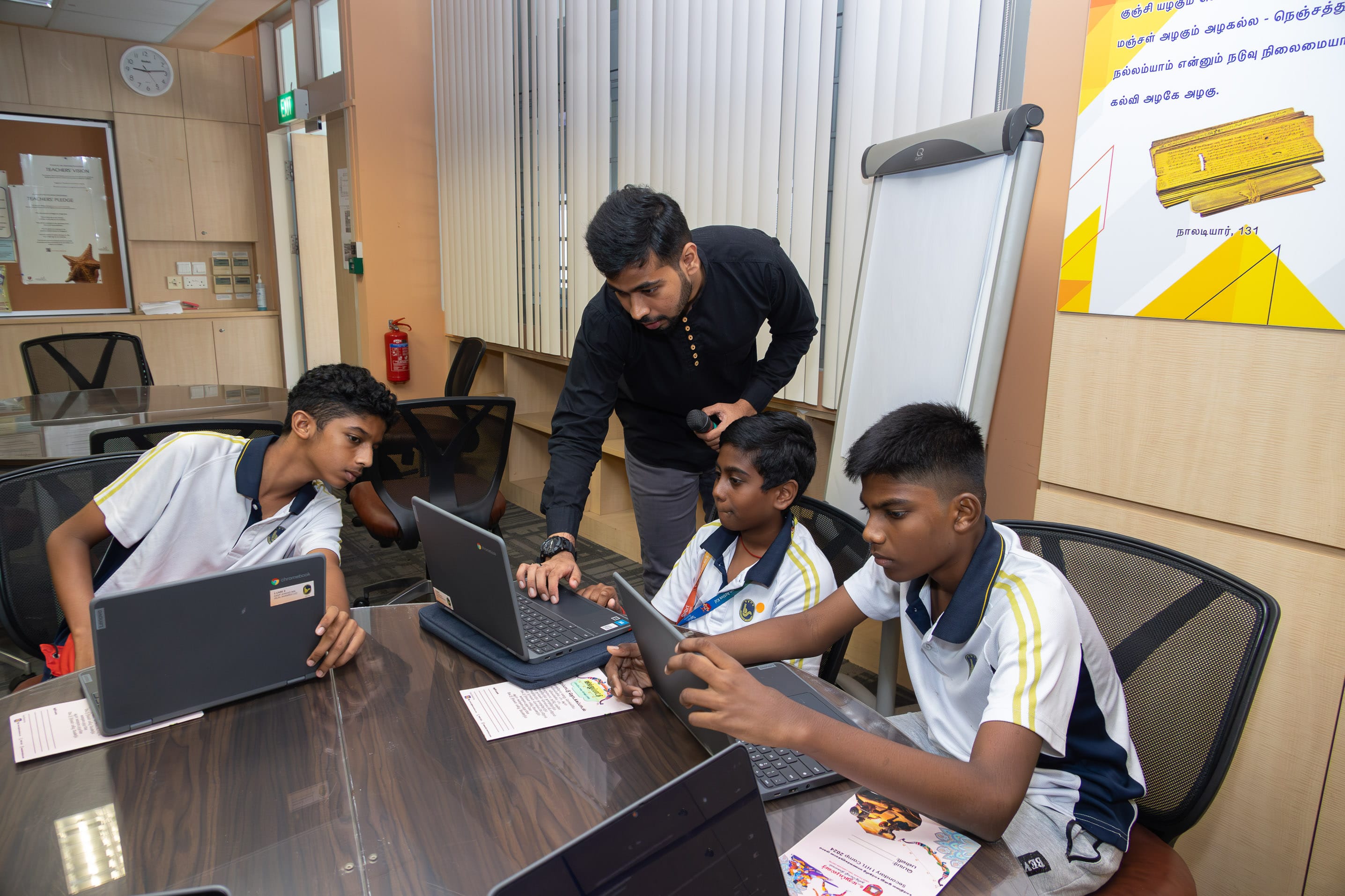 Teacher leans over table helping three students in uniform using laptops and tablets in a meeting room