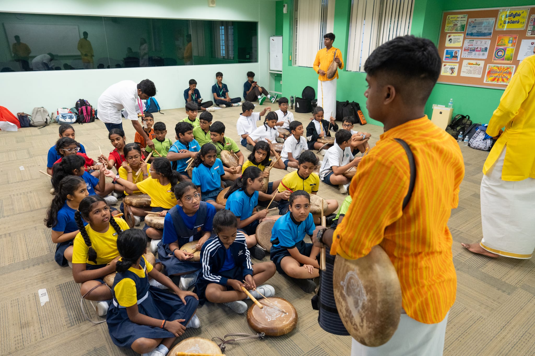 Students sit on the floor with drums as an instructor in orange stands in front, teaching rhythm.