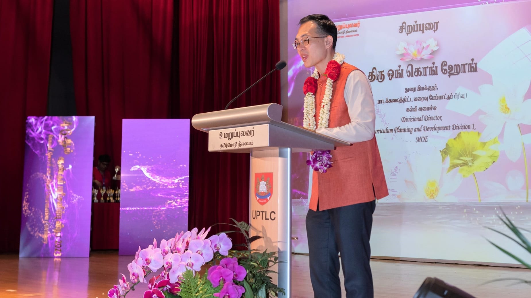 Man wearing garlands speaks at a podium on stage, with red curtains, floral backdrop, and orchids in front.