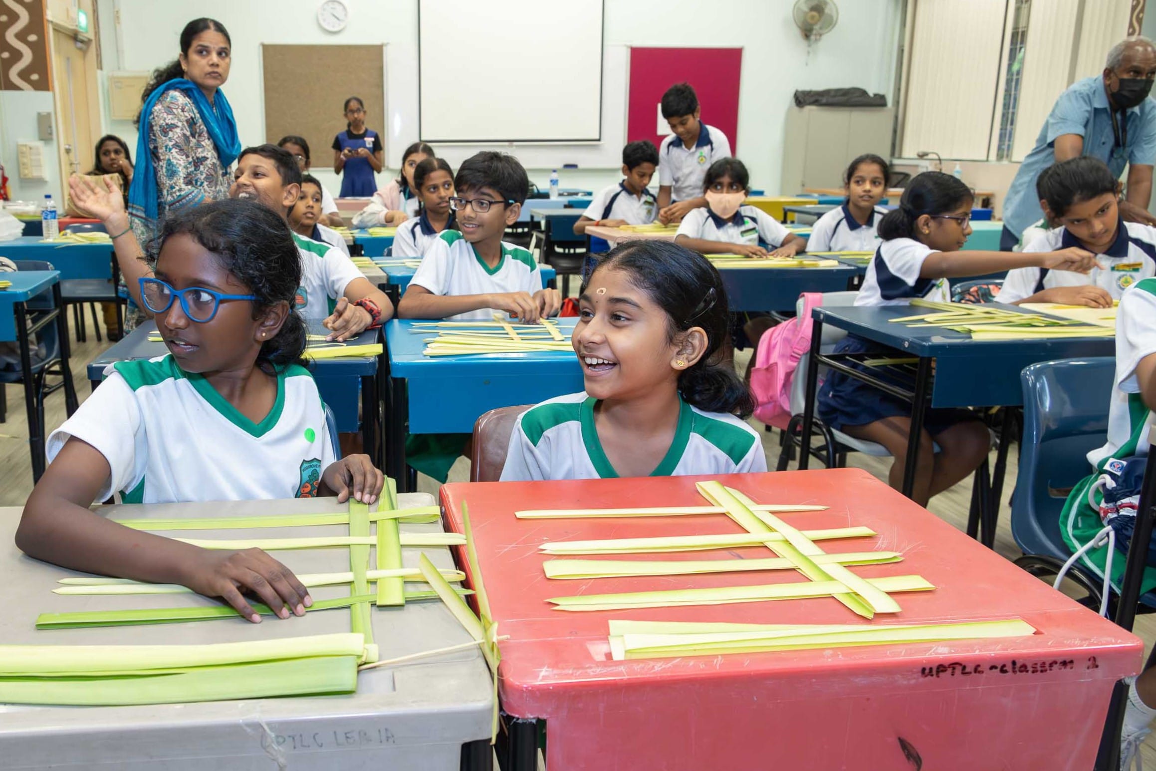 Students in a classroom weave green palm strips on desks while a teacher stands nearby.