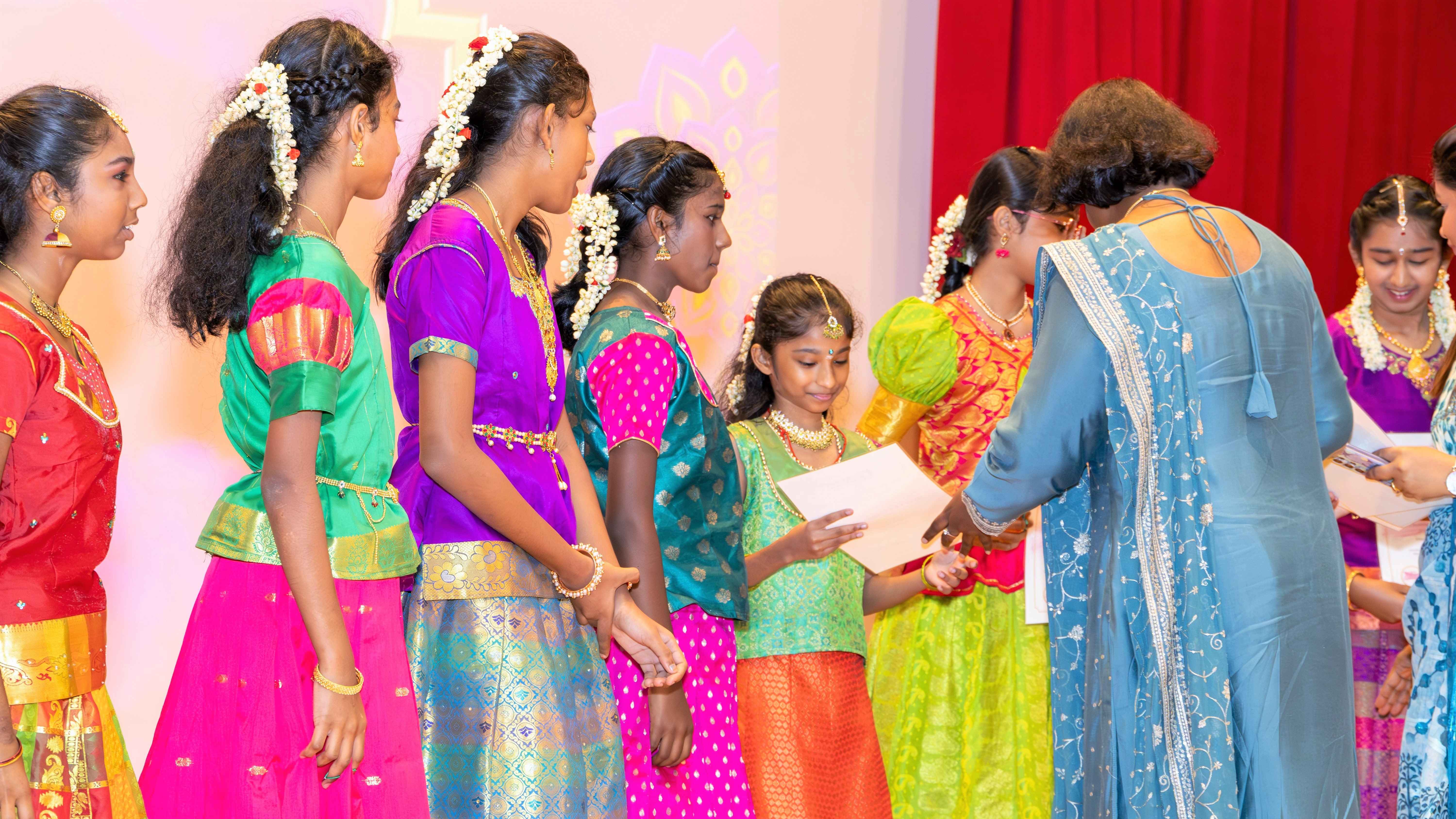 Girls in colorful dresses line up as a woman in blue hands one girl a paper on a stage with red curtain.