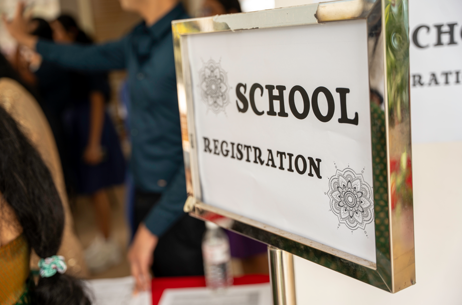 Sign reading SCHOOL REGISTRATION on a metal stand, with blurred people and a table in the background.