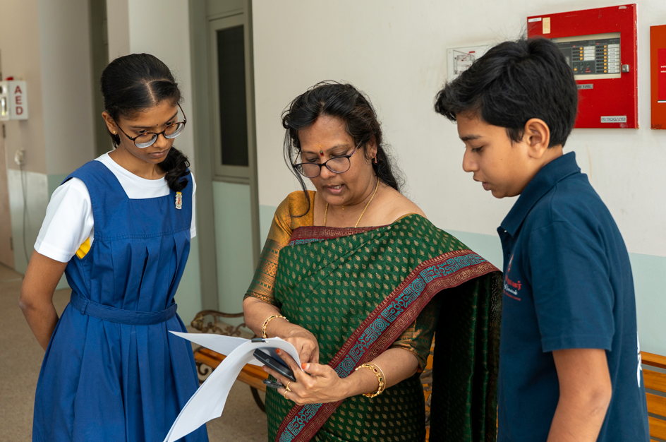 Woman in a green sari shows papers and a phone to two students in school uniforms in a hallway.
