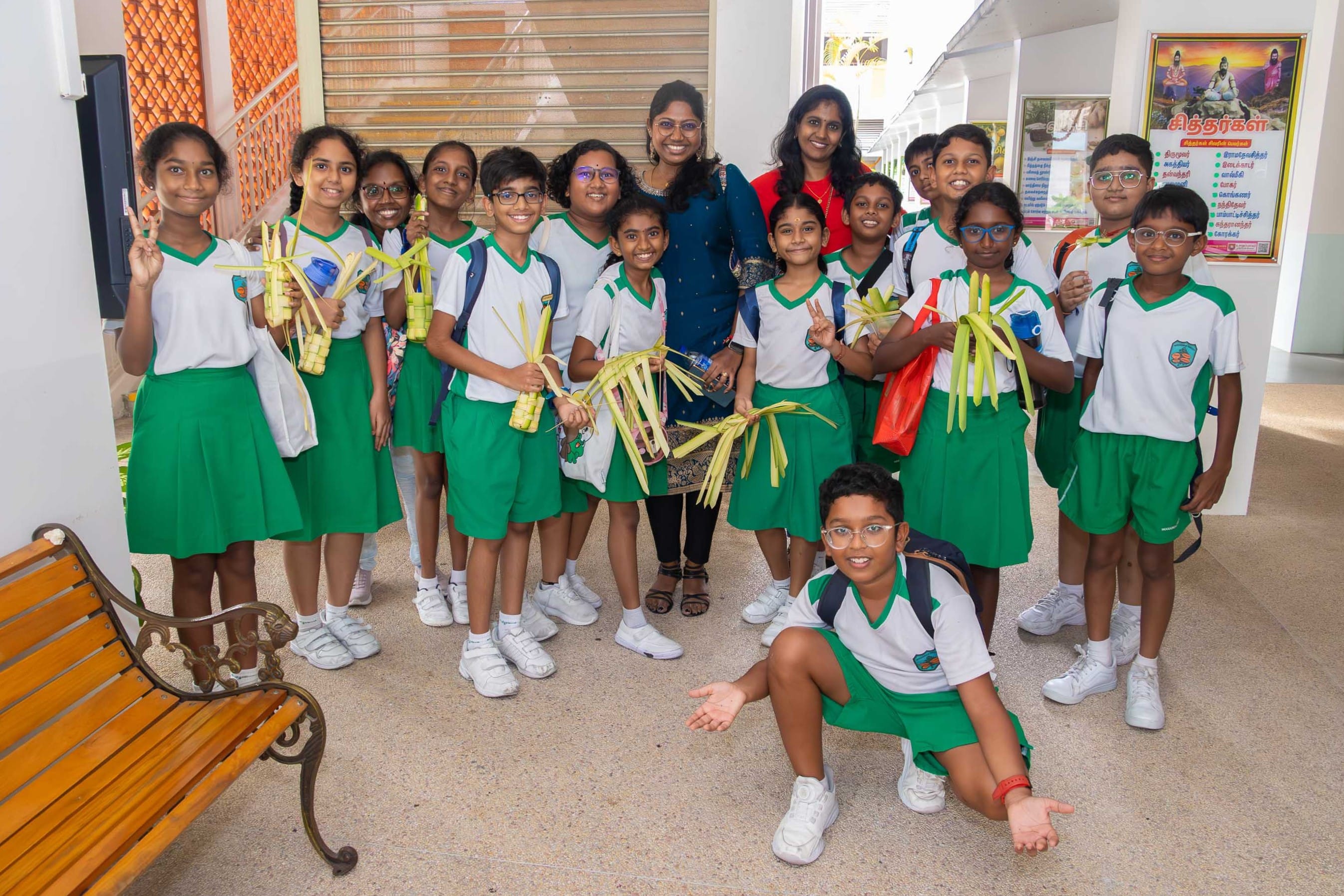 Group of schoolchildren in green uniforms with two women, holding woven palm leaves, one child crouching front.