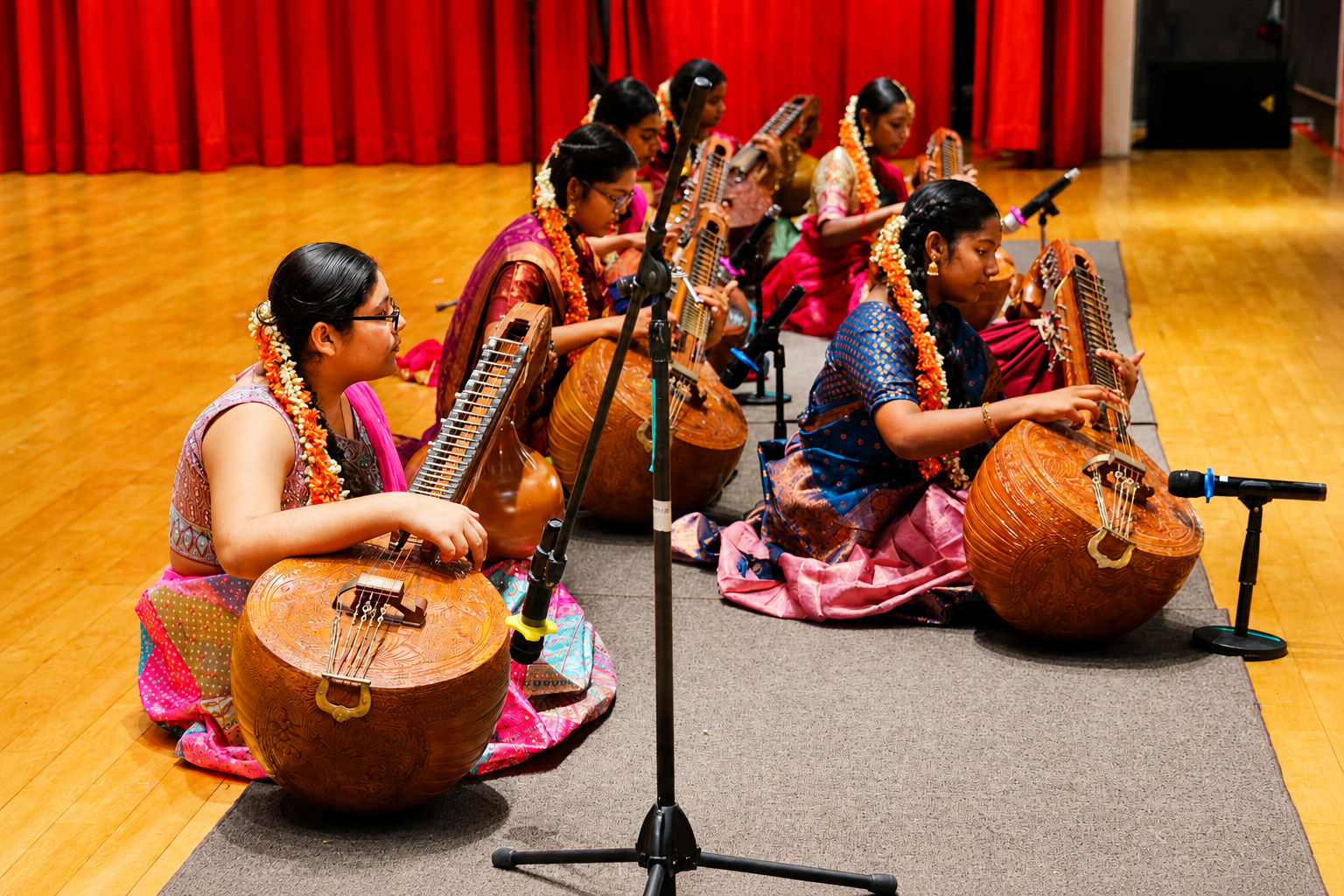 Six women in colorful saris sit on a stage playing veenas, with microphones and red curtains behind.