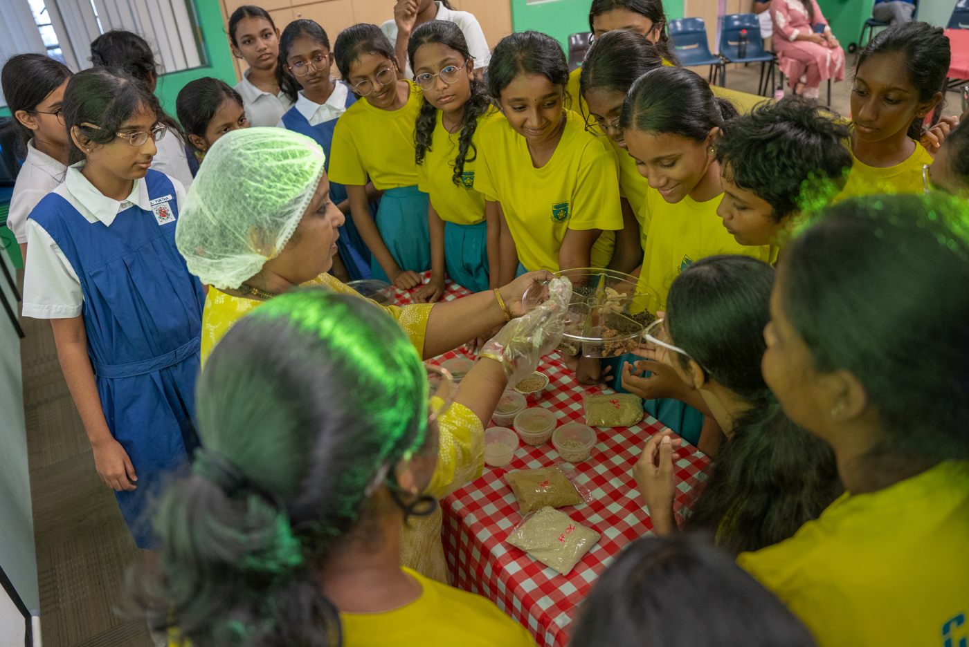 Woman in hairnet and gloves demonstrates food samples as schoolgirls gather around a checkered table.