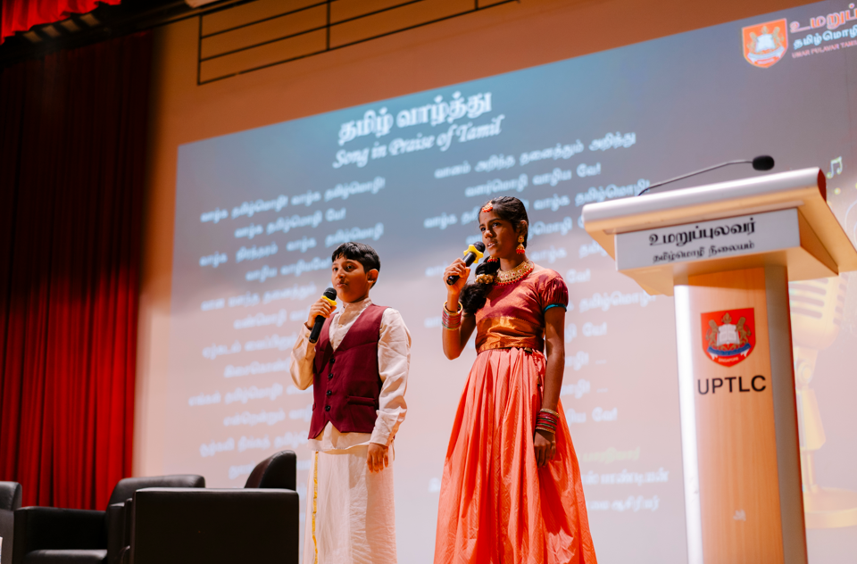 Boy and girl in traditional dress sing into microphones on stage beside a UPTLC podium.
