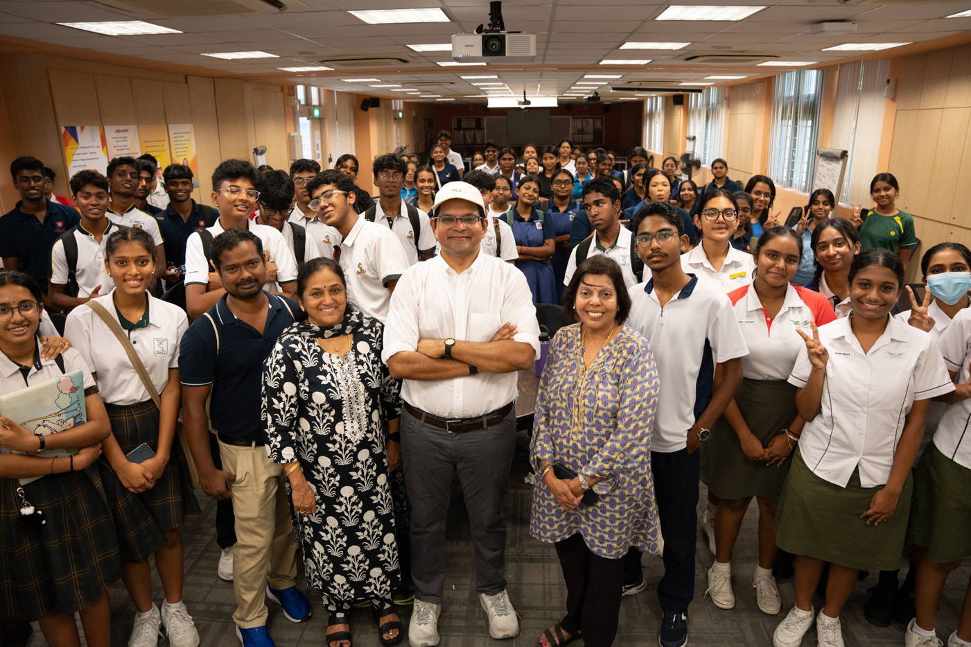 A large group of students in school uniforms and three adults pose together smiling in an indoor hall.
