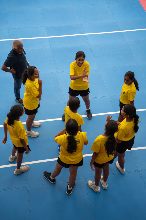 Girls in yellow shirts circle on a blue court, one clapping in center as an adult watches.