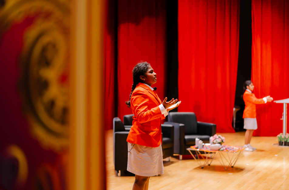 Person in orange blazer speaks onstage before red curtains; sofas and table behind, another person at podium right.