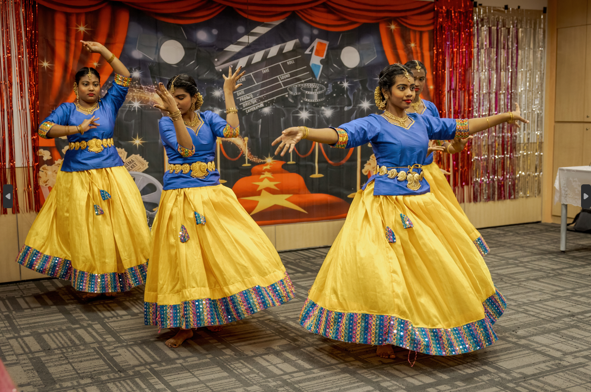 Four dancers in blue tops and yellow skirts perform on a stage with a movie-themed backdrop and red curtains.