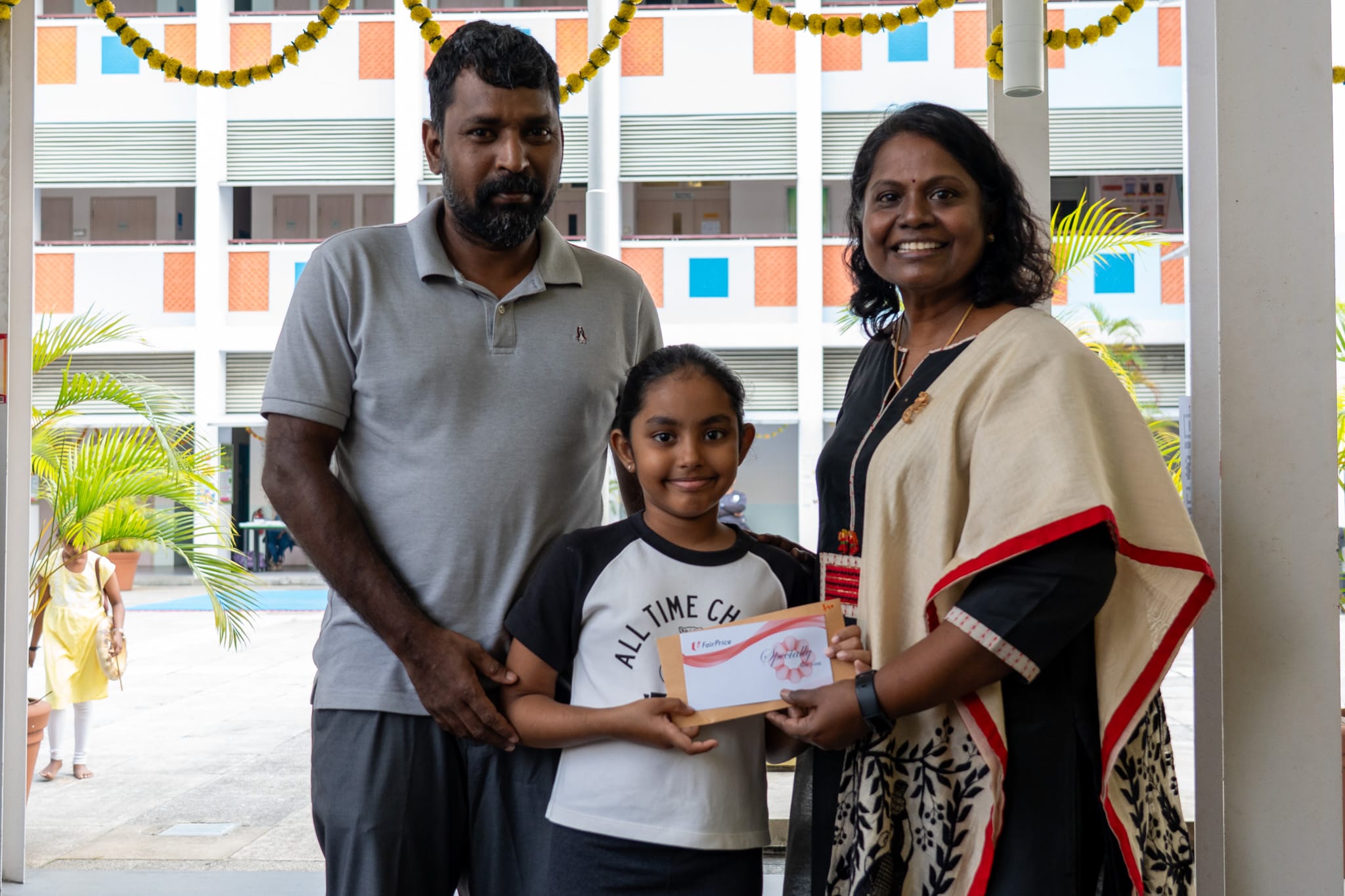 Man and woman stand beside a smiling girl holding a certificate envelope in a school courtyard.