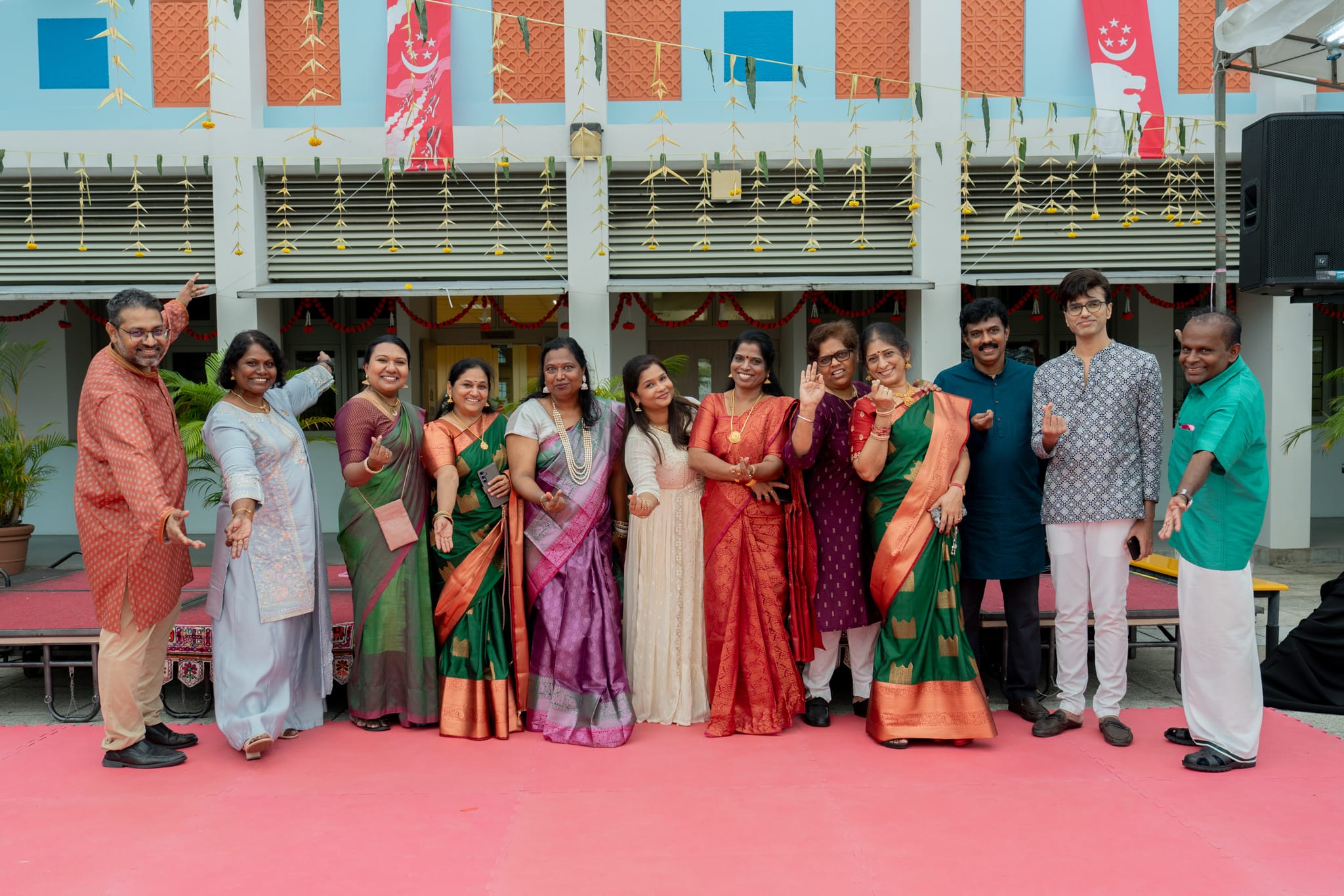 A group of people in colorful traditional Indian attire pose on a pink stage with festive decorations behind them.