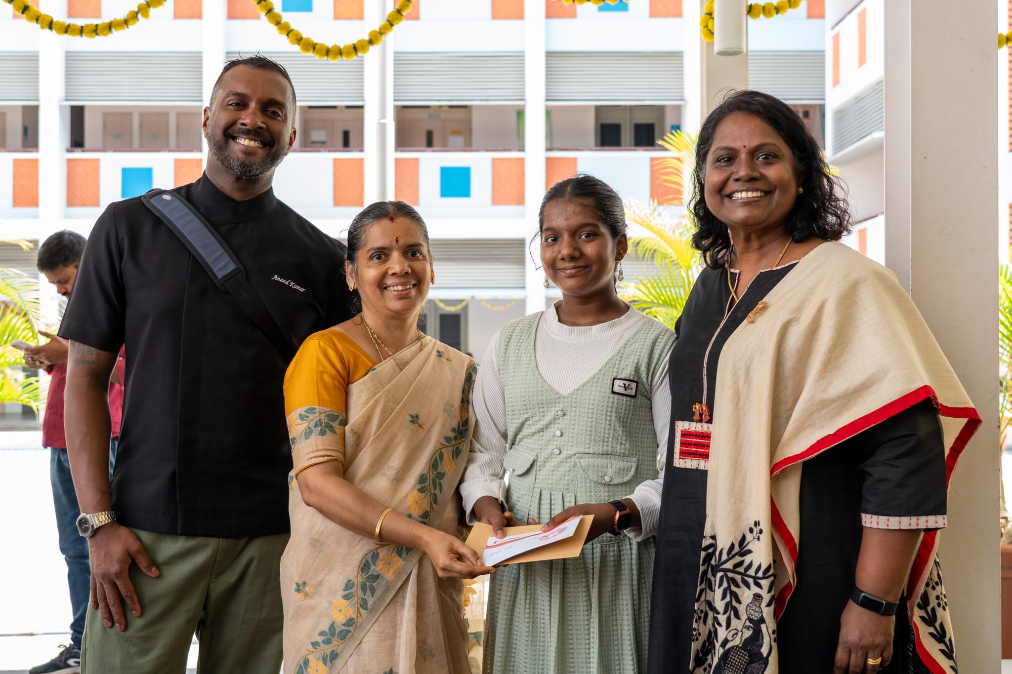 Four people smiling as a woman hands a girl an envelope in a courtyard decorated with garlands.
