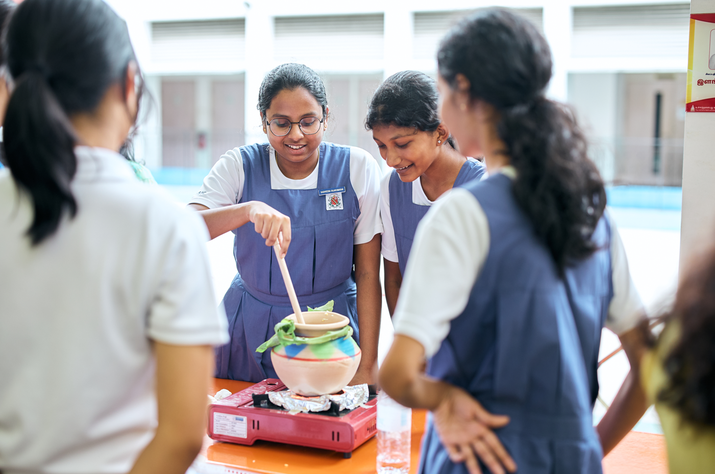 Students in school uniforms gather around a table, stirring a pot on a red portable stove.