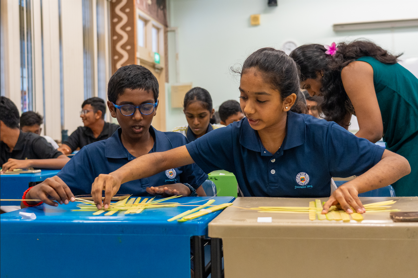 Students in navy uniforms weave pale strips on desks in a classroom as a teacher leans over to help.
