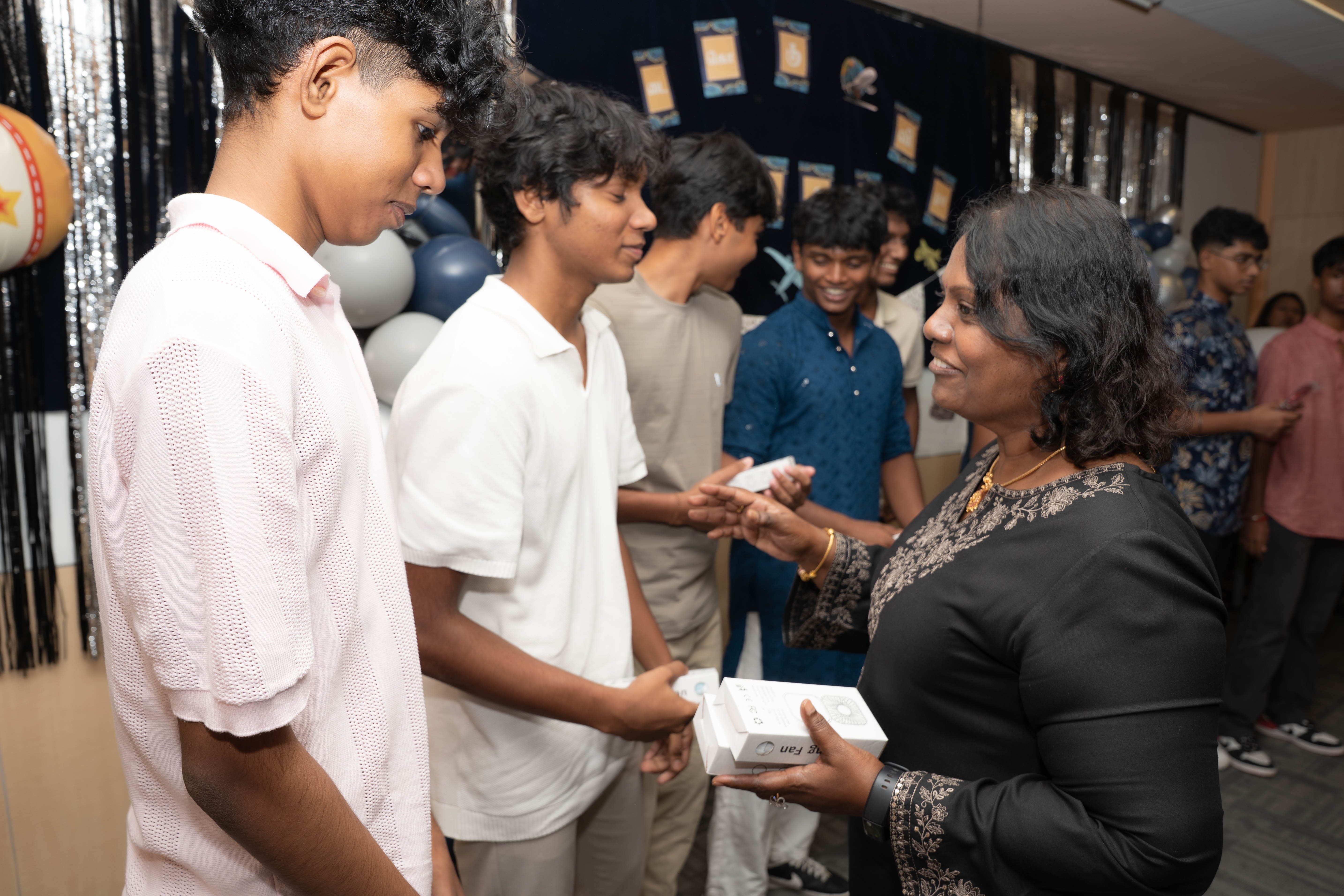 Woman holding a box speaks with two young men as others smile in a decorated room with balloons.