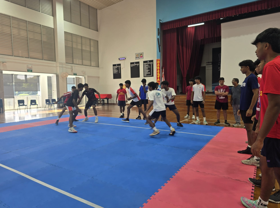 Students play a team sport on blue mats in a gym, with others watching near a stage with red curtains.