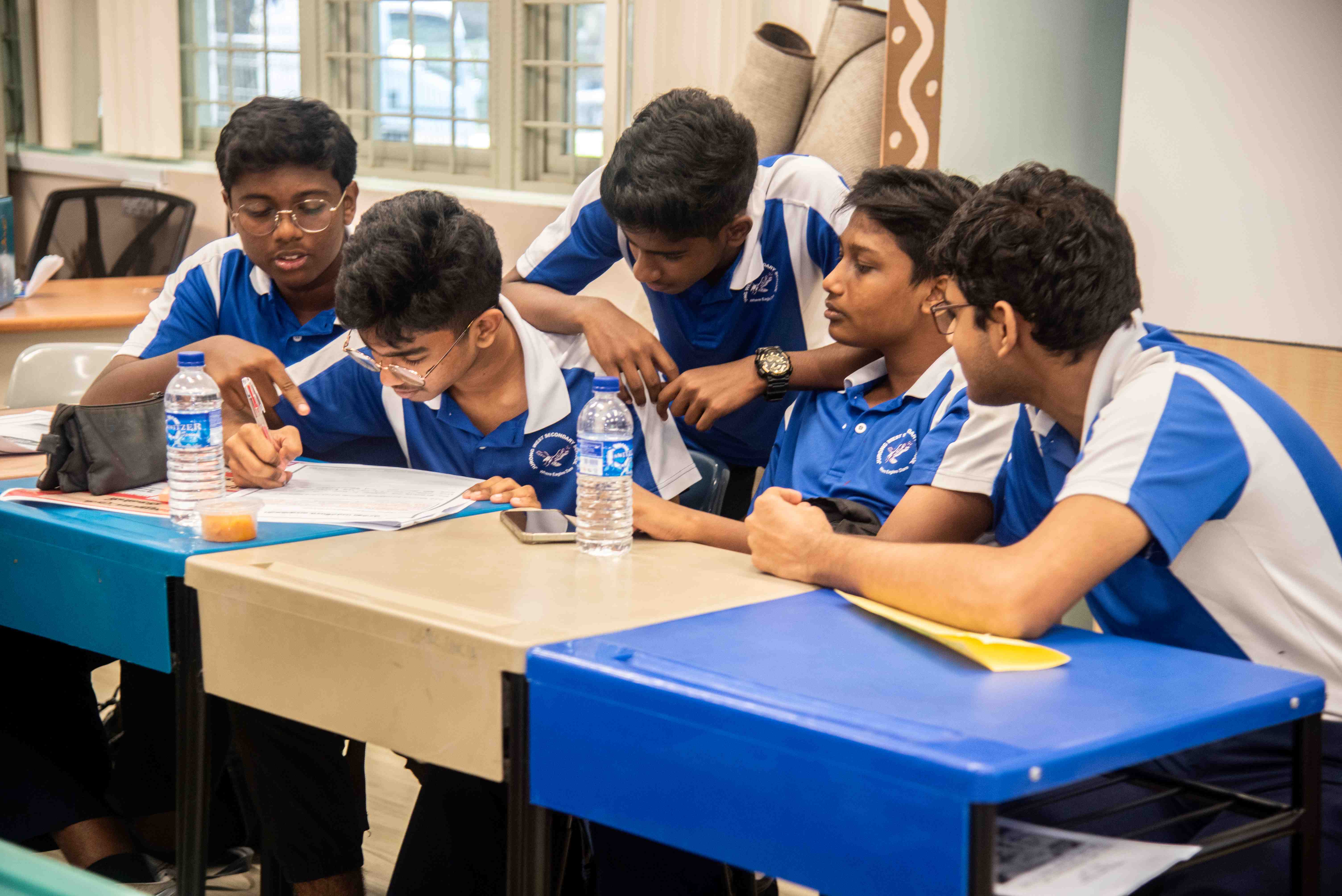 Five students in blue uniforms sit at desks, one writes on paper while others look on.
