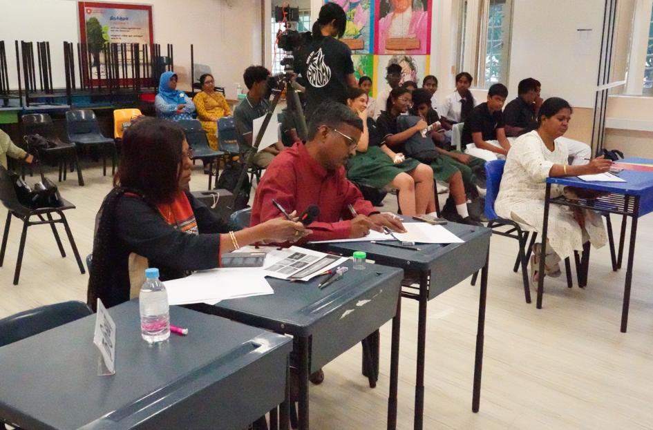 Two adults write at desks while students sit behind them; a camera on a tripod stands in the room.