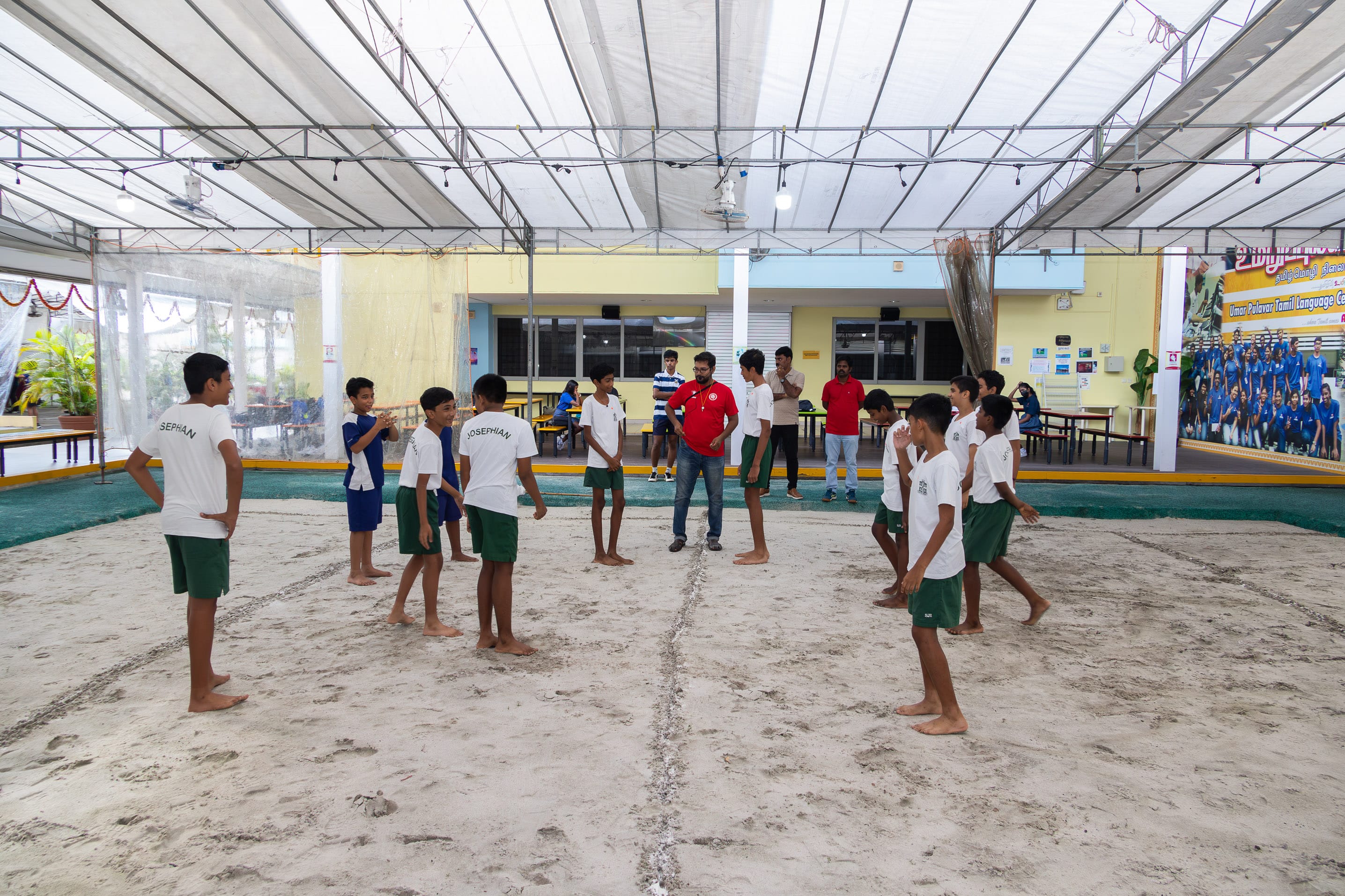 Boys in white shirts and green shorts stand barefoot on a sand court under a canopy, with coaches behind.