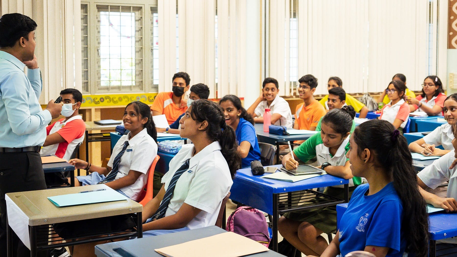 Teacher speaking to a classroom of students at desks; some smile and one writes in a notebook.