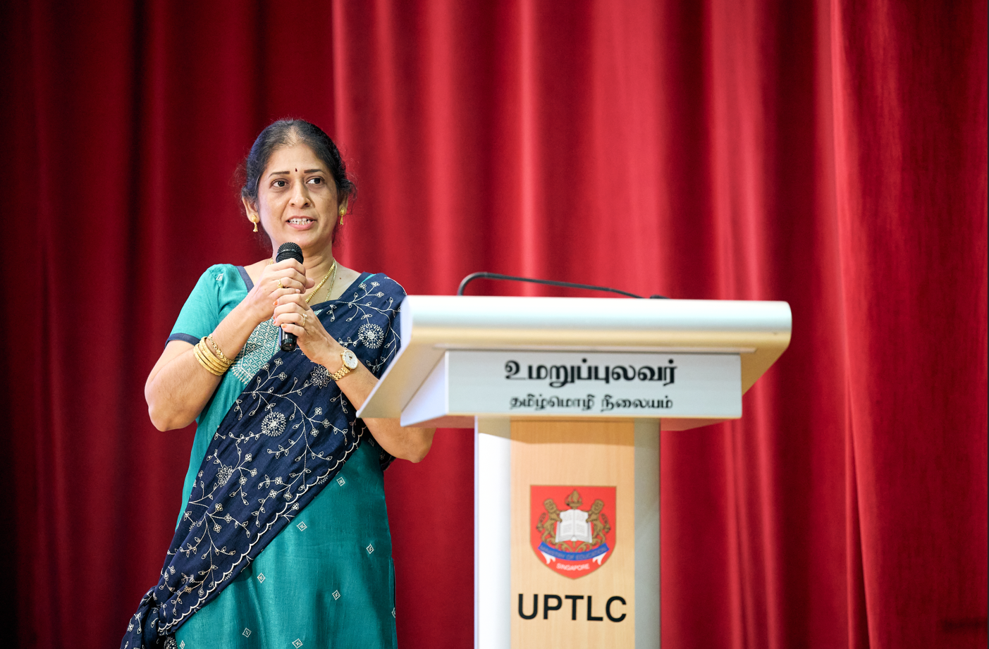 Woman in sari speaks into a microphone beside a lectern labeled UPTLC, in front of red curtains.